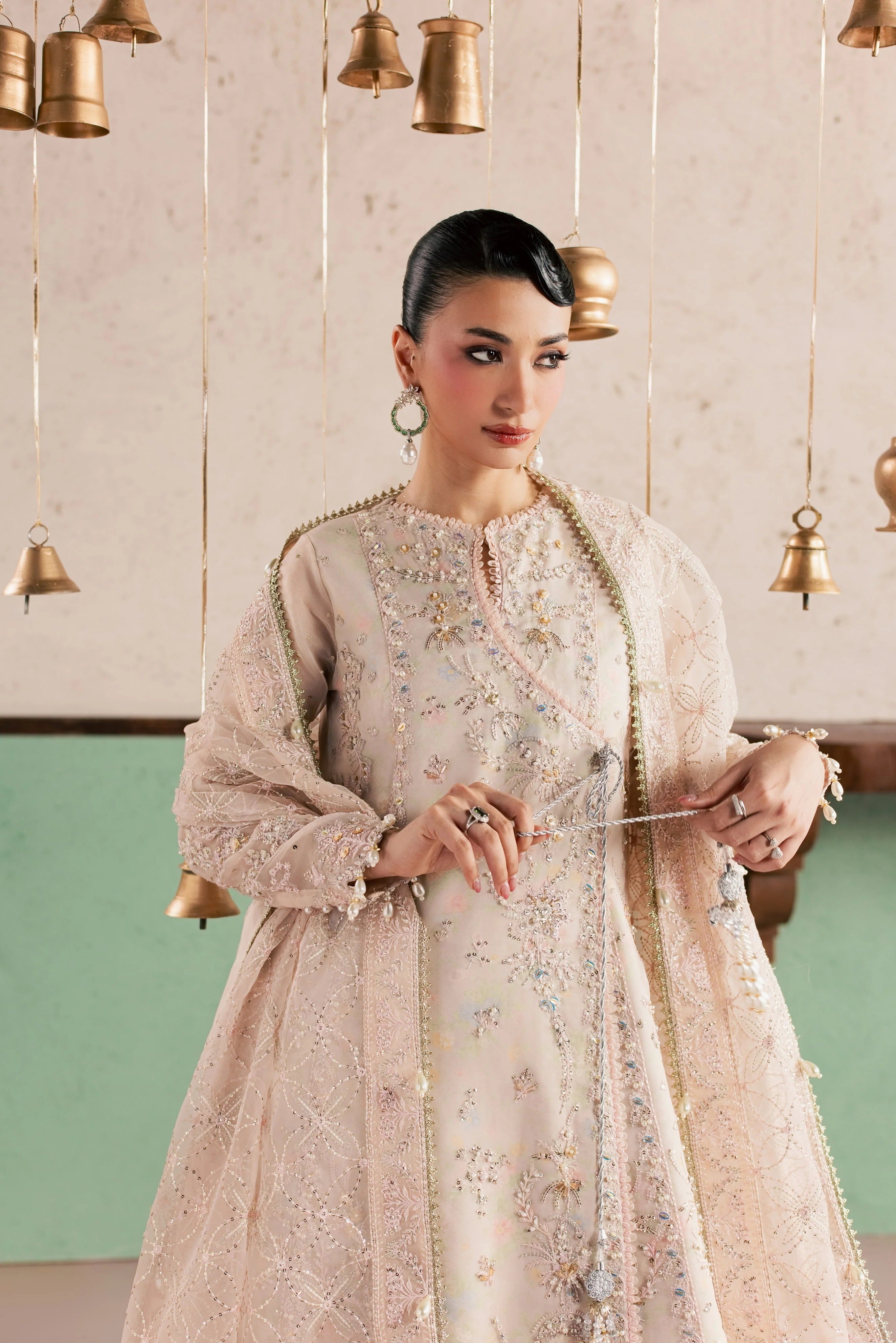 A woman models a traditional embroidered Anarkali gown in a light beige or cream color with intricate gold and multicolor floral embroidery. She is standing in front of a decorative wall with hanging brass bells.