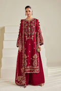 A woman models a deep red embroidered traditional outfit with gold detailing, consisting of a long kurta, matching pants, and sheer sleeves, set against a minimalist beige background with white steps.