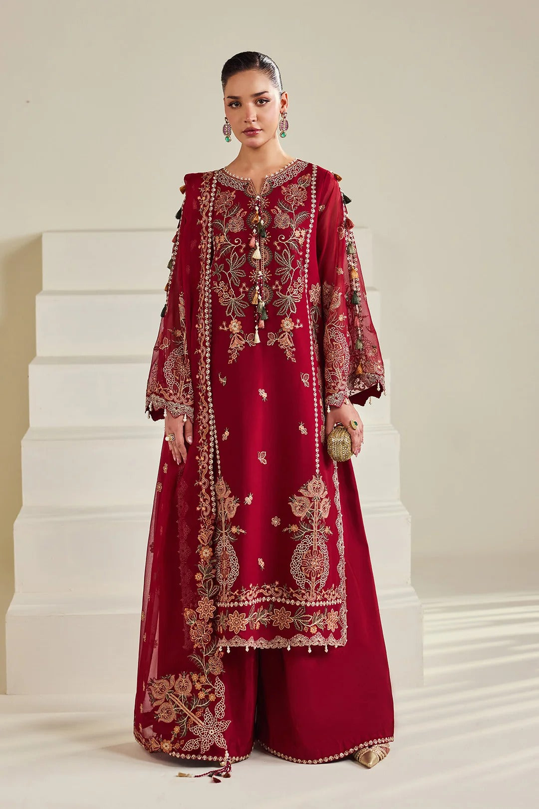 A woman models a deep red embroidered traditional outfit with gold detailing, consisting of a long kurta, matching pants, and sheer sleeves, set against a minimalist beige background with white steps.