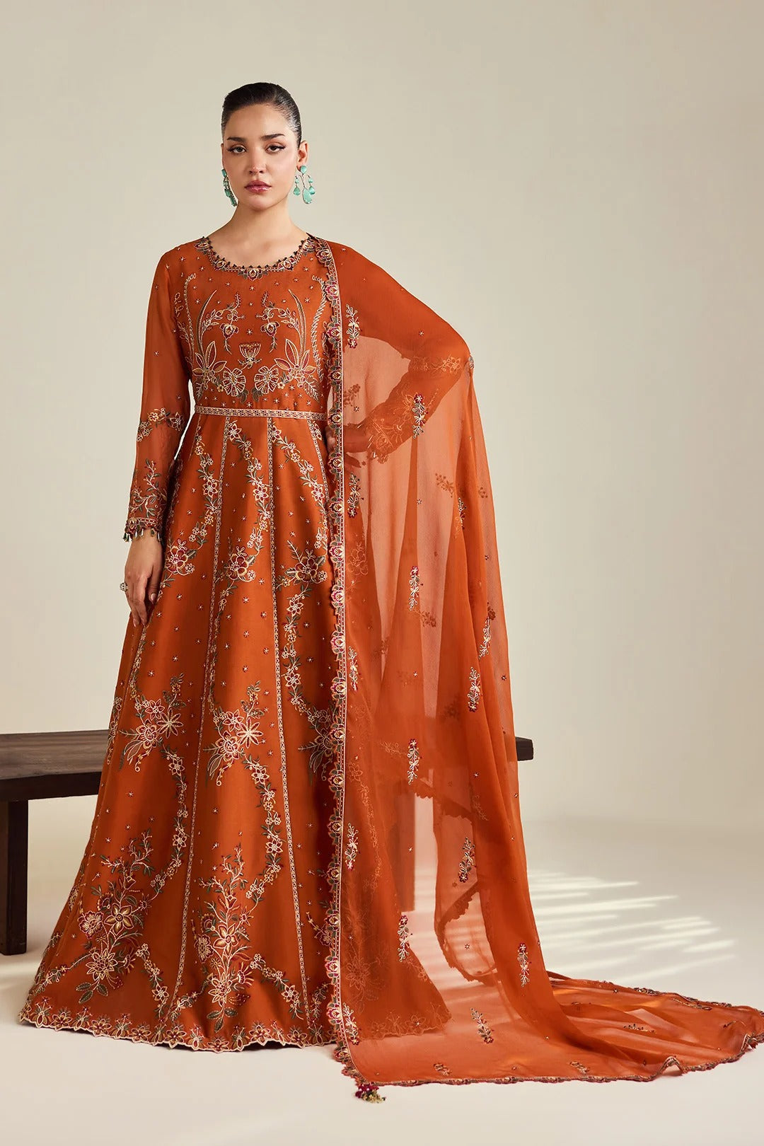 A woman models a rust-orange embroidered Anarkali gown with a matching sheer dupatta, set against a neutral beige studio background.