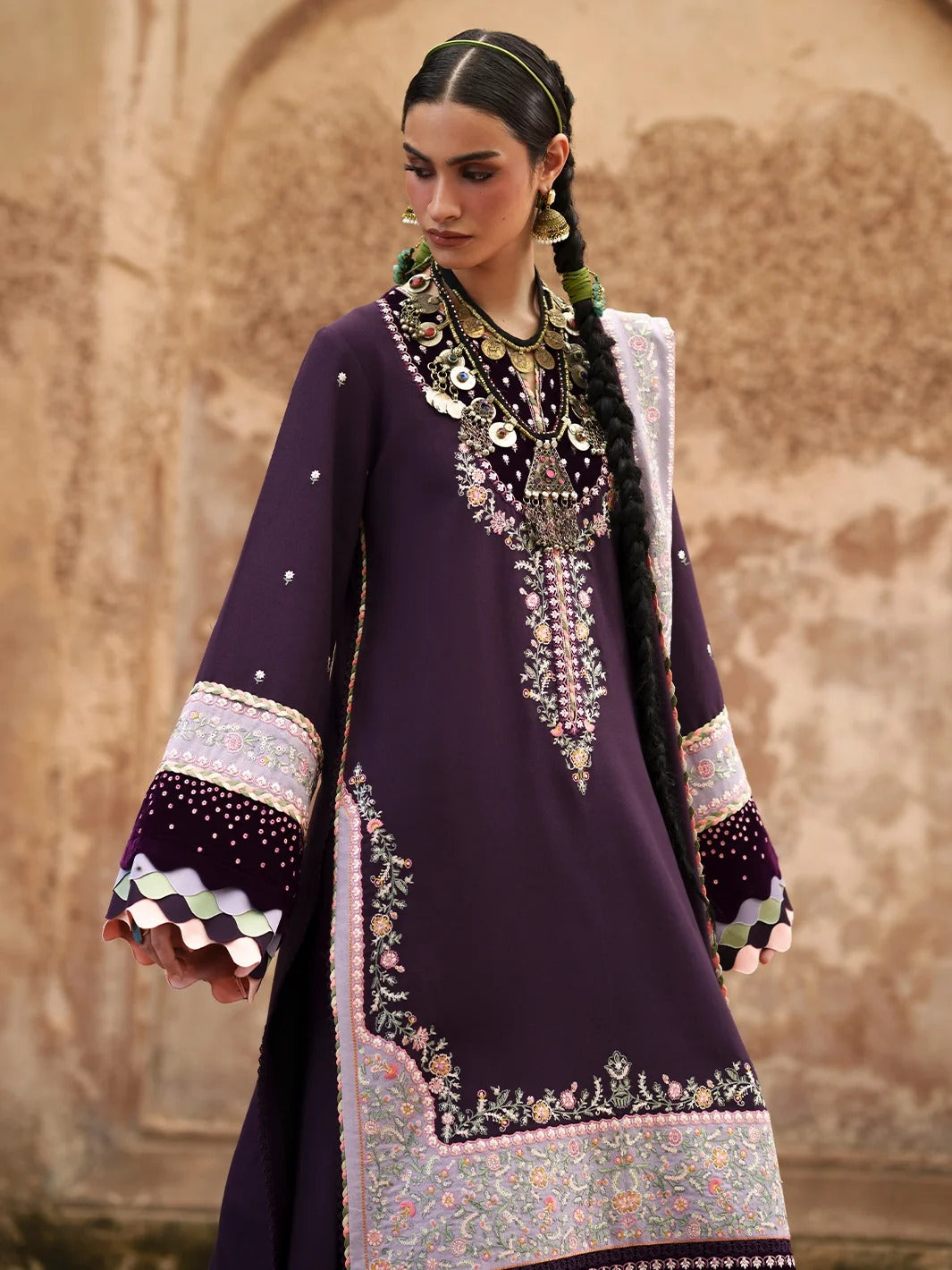 A woman modeling a deep purple traditional South Asian outfit with intricate silver and multicolor embroidery, layered with heavy gold necklaces, against a warm beige stone wall background.