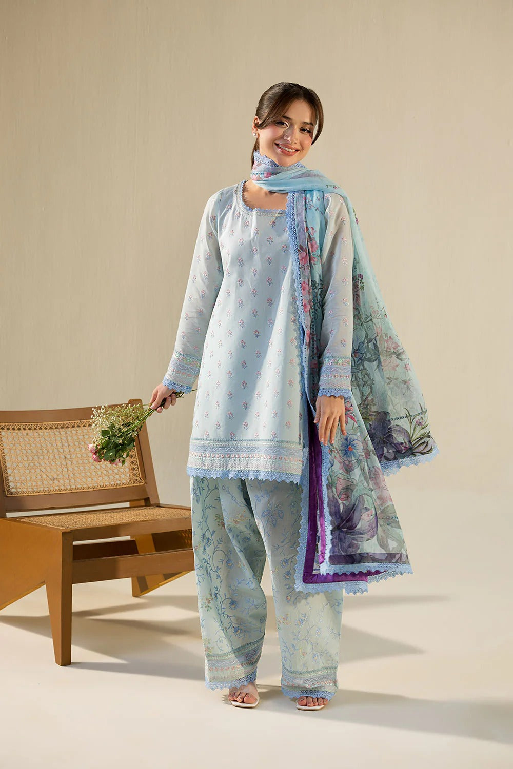 Woman in a light blue traditional outfit with a matching dupatta, holding a basket, on a beige background.