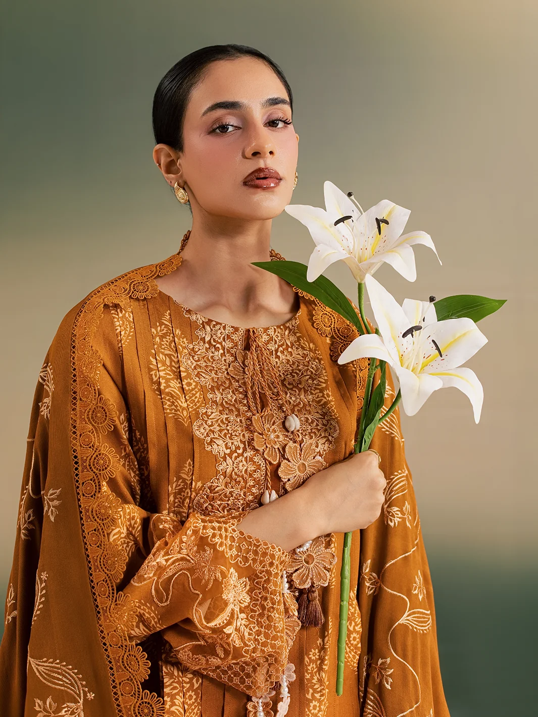 A woman wearing a golden-brown embroidered traditional outfit with a matching draped shawl, holding two white lilies against a soft beige background.