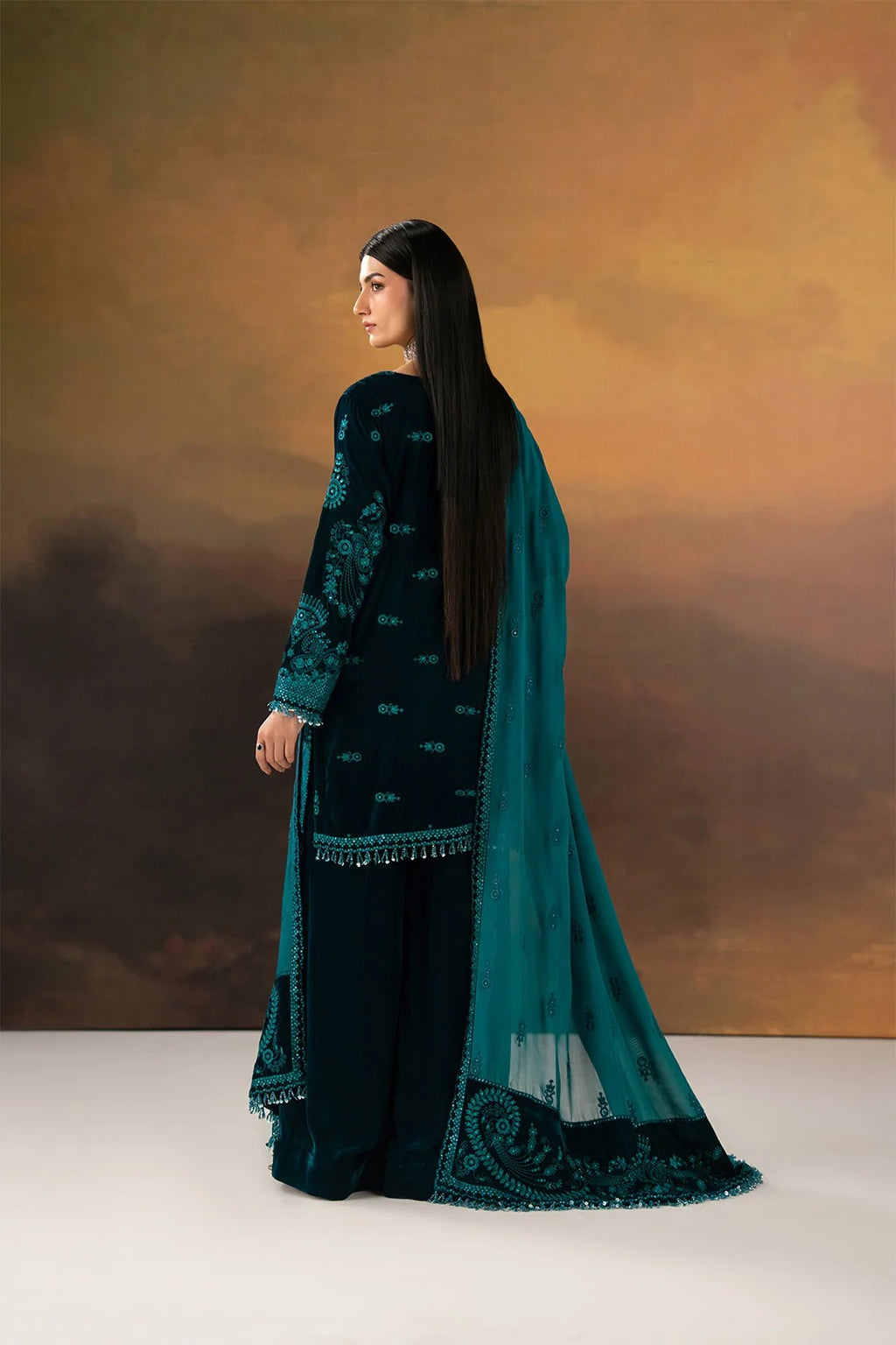 A woman modeling a dark teal and black traditional South Asian outfit with intricate embroidery and a matching sheer dupatta, posed against a warm brown and gold studio backdrop.