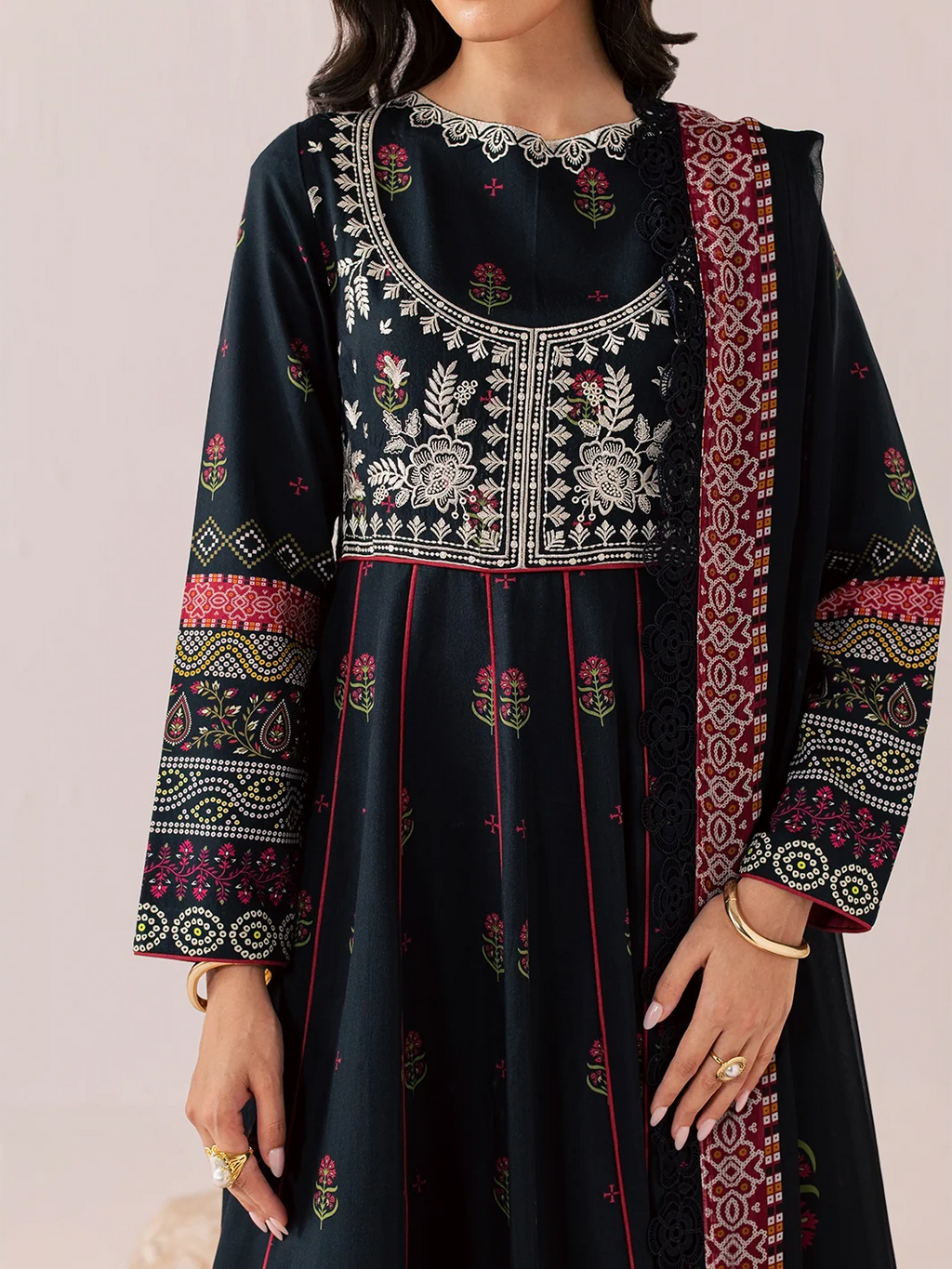 A woman wearing a dark navy or black embroidered kurta with intricate white, red, and green floral patterns, paired with a matching patterned dupatta, set against a soft beige background.