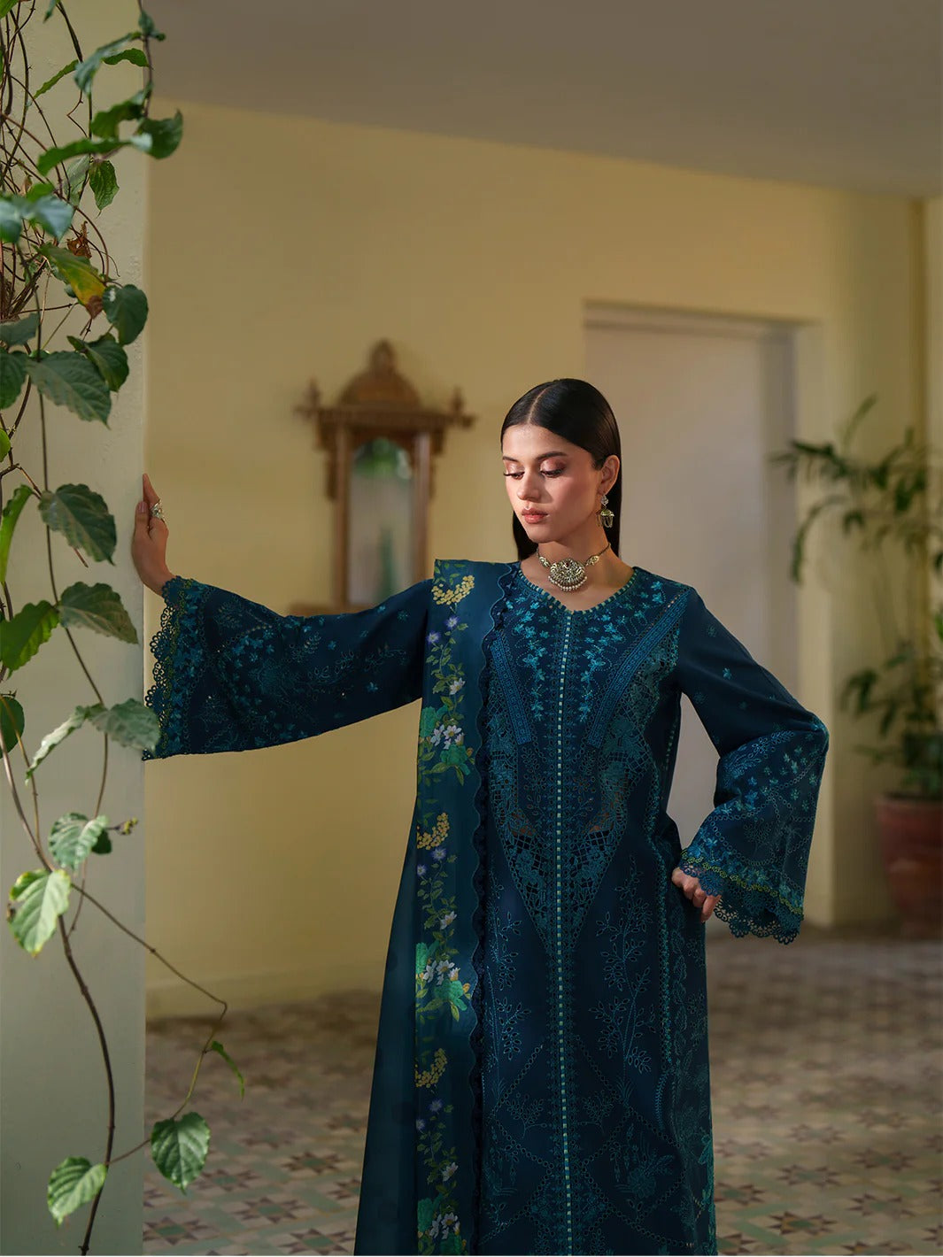 Woman in a teal embroidered dress standing indoors with plants around
