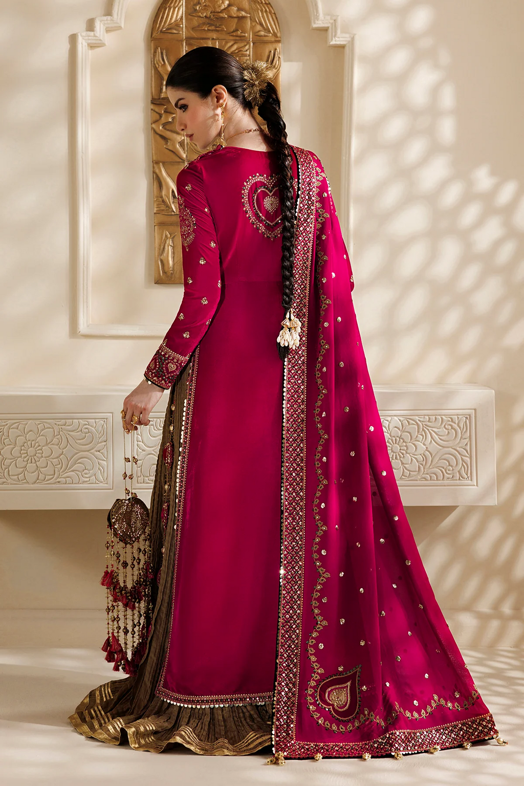 A woman modeling a traditional Indian salwar kameez outfit in a warm beige interior. The outfit consists of a deep magenta-pink kurta with gold embroidery, a matching dupatta, and a brown embroidered border.