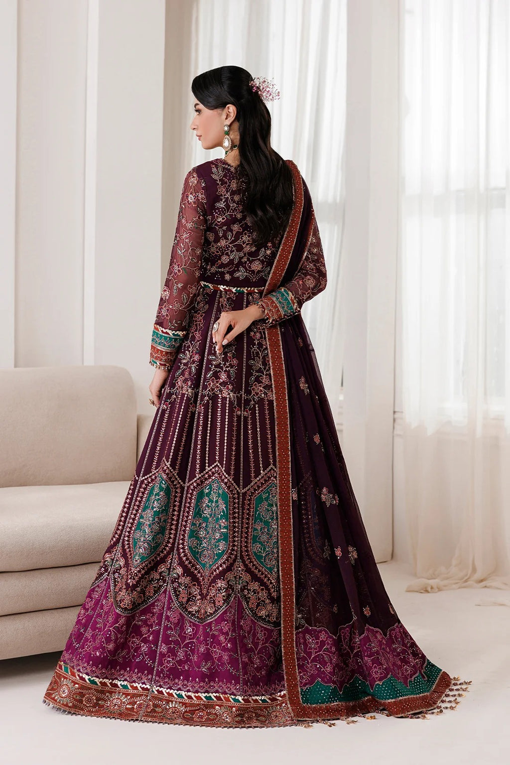 A woman modeling a traditional Indian anarkali gown in deep purple and magenta with intricate gold embroidery and teal accents, photographed in a bright indoor setting with a sofa and sheer curtains.