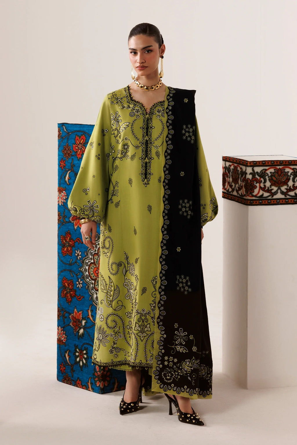 A woman models a traditional long kurta in olive green and black with intricate gold embroidery, standing in a studio setting with decorative patterned panels in the background.