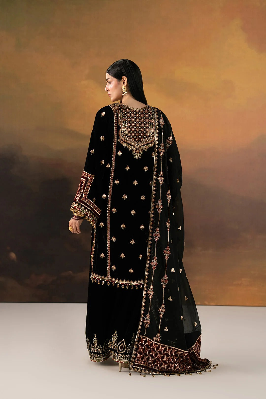 A woman modeling a black traditional embroidered outfit with ornate gold and brown detailing, shown from the back against a warm brown studio backdrop.