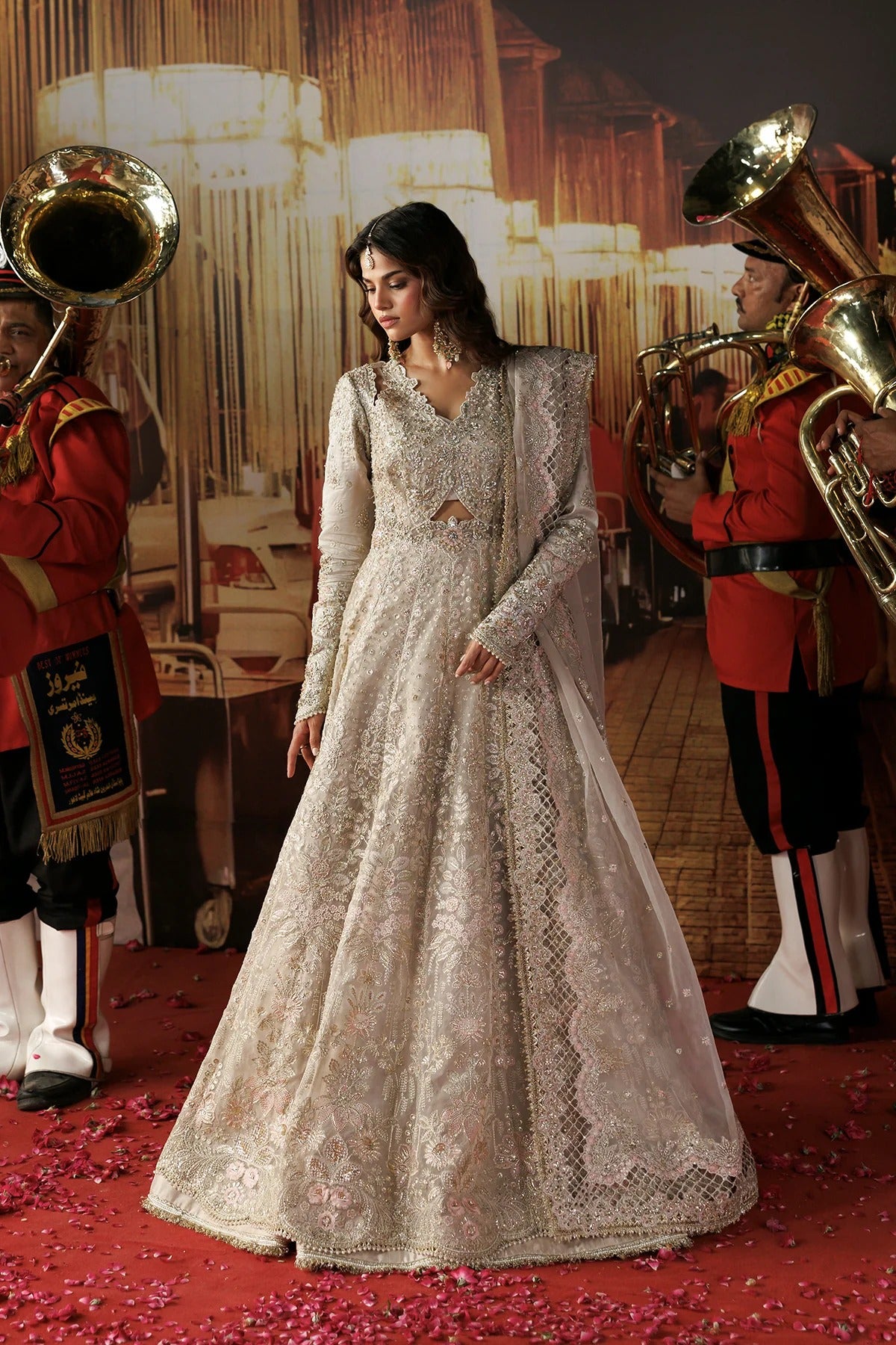 A woman wearing a heavily embroidered off-white/cream traditional gown with long sleeves and a flared skirt, standing on a red carpet with red flower petals. Two uniformed brass instrument players in red jackets are visible on both sides against a warm-toned stage backdrop.
