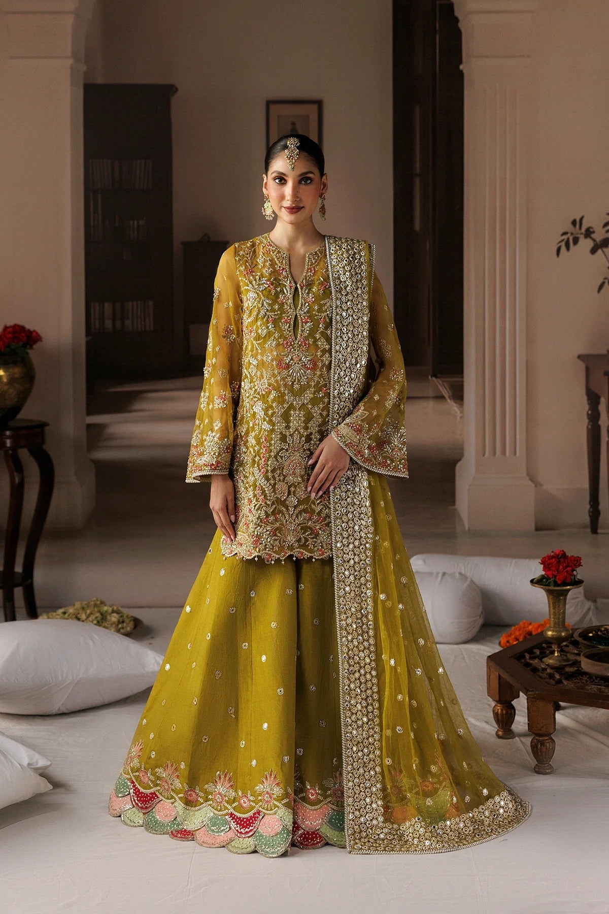 A woman models a traditional Indian ensemble featuring a mustard yellow embroidered kurta and flared pants with a matching sheer dupatta, set in an elegant indoor room.