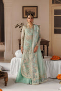 A woman stands indoors wearing an ornate mint green traditional outfit with heavy beige and gold embroidery, a matching embroidered outer layer, and a sheer dupatta. The setting is a decorated room with white draped furniture, framed wall art, and orange flowers on tables.