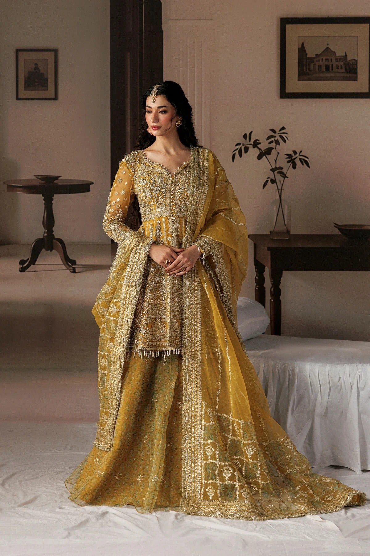 A woman models a traditional Indian ensemble consisting of a heavily embroidered mustard yellow kurta, matching flared skirt, and dupatta, set against a softly lit, vintage-style indoor room background.