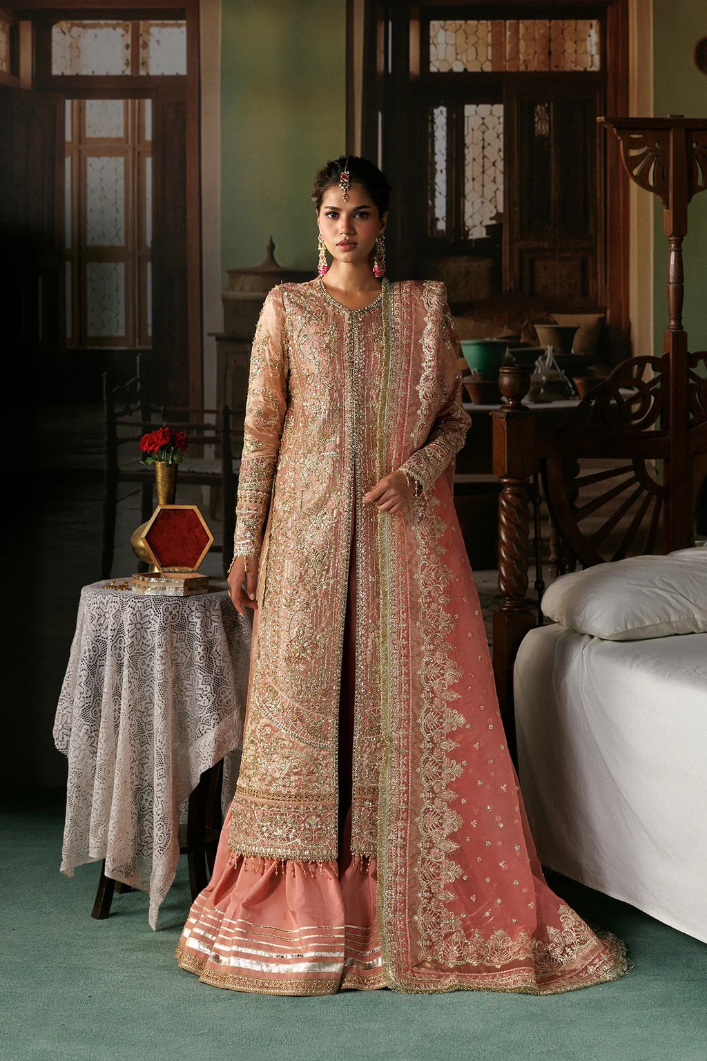 A woman models a traditional Indian salwar kameez outfit in a rustic indoor setting. The outfit features a peach-pink dupatta and flared skirt with intricate gold embroidery, paired with a matching beige and gold embroidered long kurta.