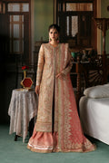 A woman models a traditional Indian salwar kameez outfit in a rustic indoor setting. The outfit features a peach-pink dupatta and flared skirt with intricate gold embroidery, paired with a matching beige and gold embroidered long kurta.