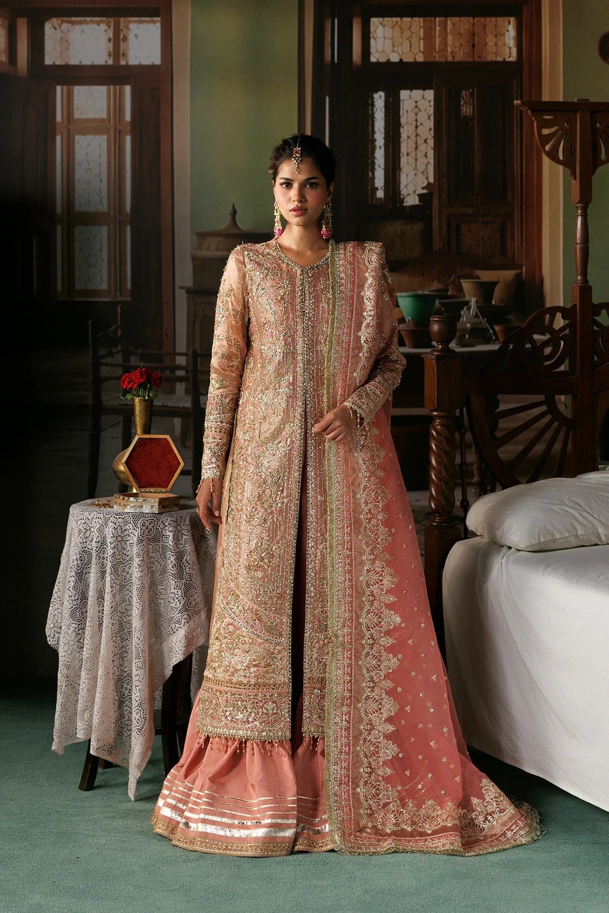 A woman models a traditional Indian salwar kameez outfit in a rustic indoor setting. The outfit features a peach-pink dupatta and flared skirt with intricate gold embroidery, paired with a matching beige and gold embroidered long kurta.