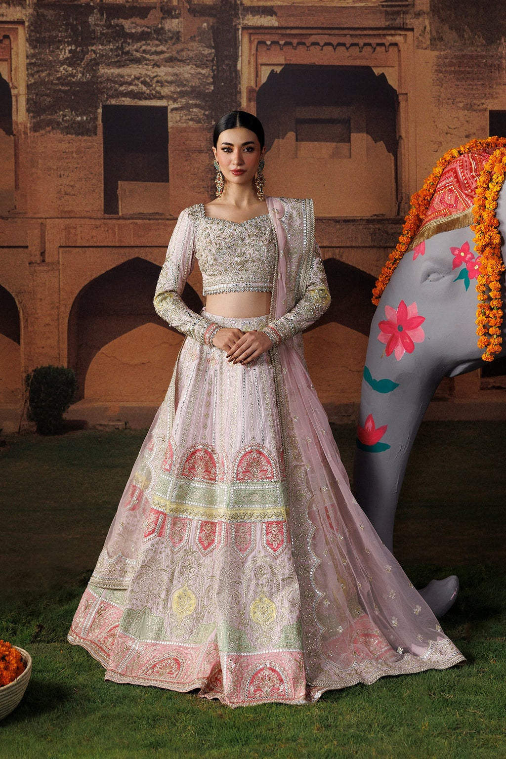 A woman models a traditional Indian lehenga choli outfit in a courtyard setting. The outfit is light pink with intricate silver embroidery and multicolor motifs including green and yellow.