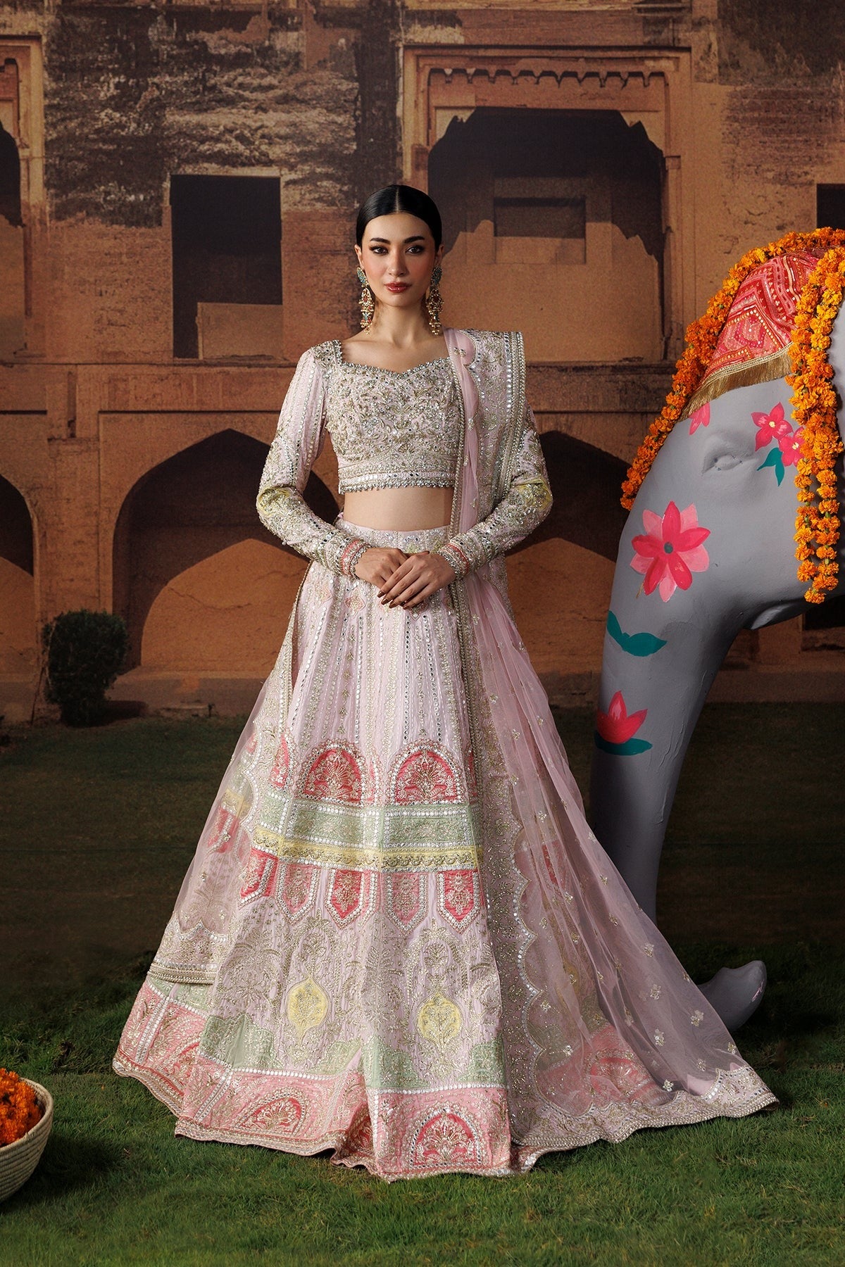 A woman models a traditional Indian lehenga choli outfit in a courtyard setting. The outfit is light pink with intricate silver embroidery and multicolor motifs including green and yellow.