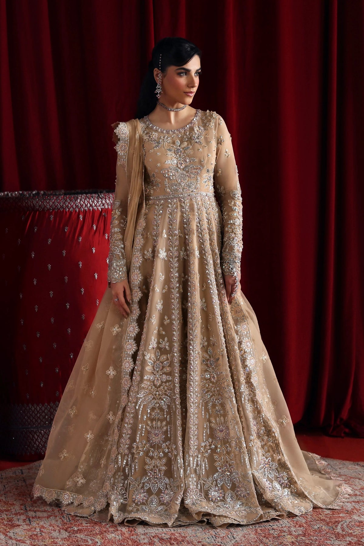 A woman models a heavily embellished beige and gold traditional gown with intricate silver embroidery, set against a dark red curtain backdrop.