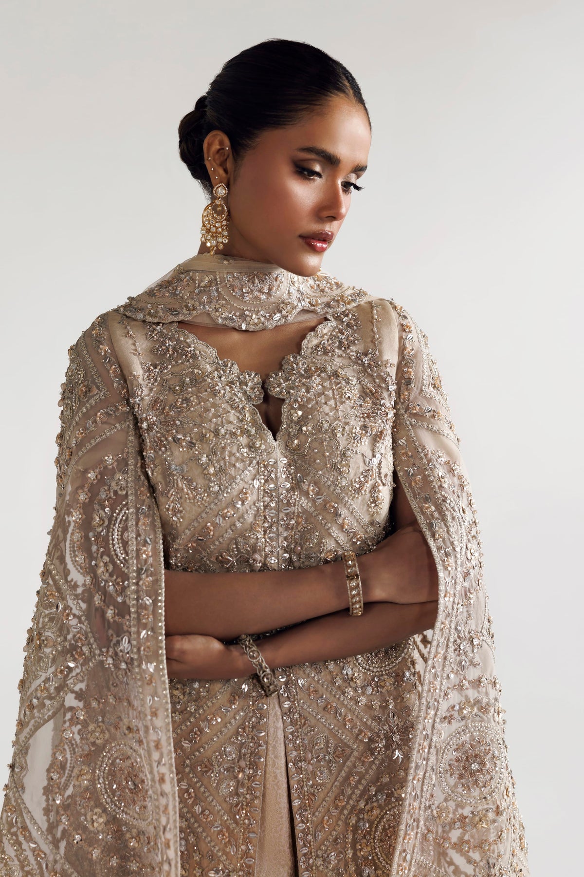 A woman wearing an ornate beige and gold embellished traditional outfit with a matching sheer embroidered cape, photographed against a plain light background.