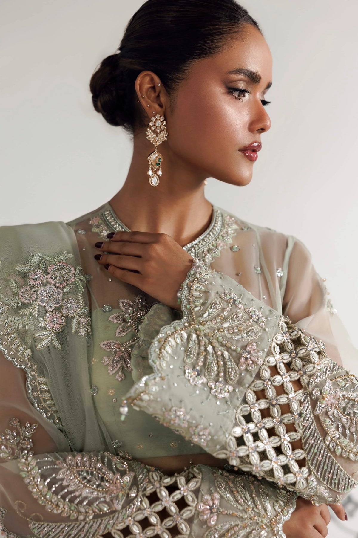 A woman in profile wearing an ornate light sage green embellished traditional outfit with intricate silver and gold embroidery, sheer sleeves, and matching dangling earrings against a plain light background.