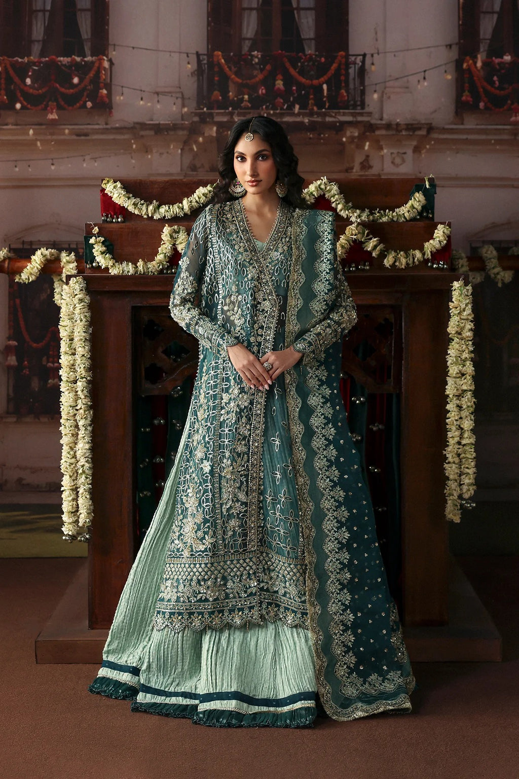 A woman stands indoors wearing an ornate teal and mint green traditional outfit with heavy gold embroidery, a long sheer jacket, flared skirt, and matching dupatta. The setting features a wooden structure decorated with floral garlands and warm ambient lighting.
