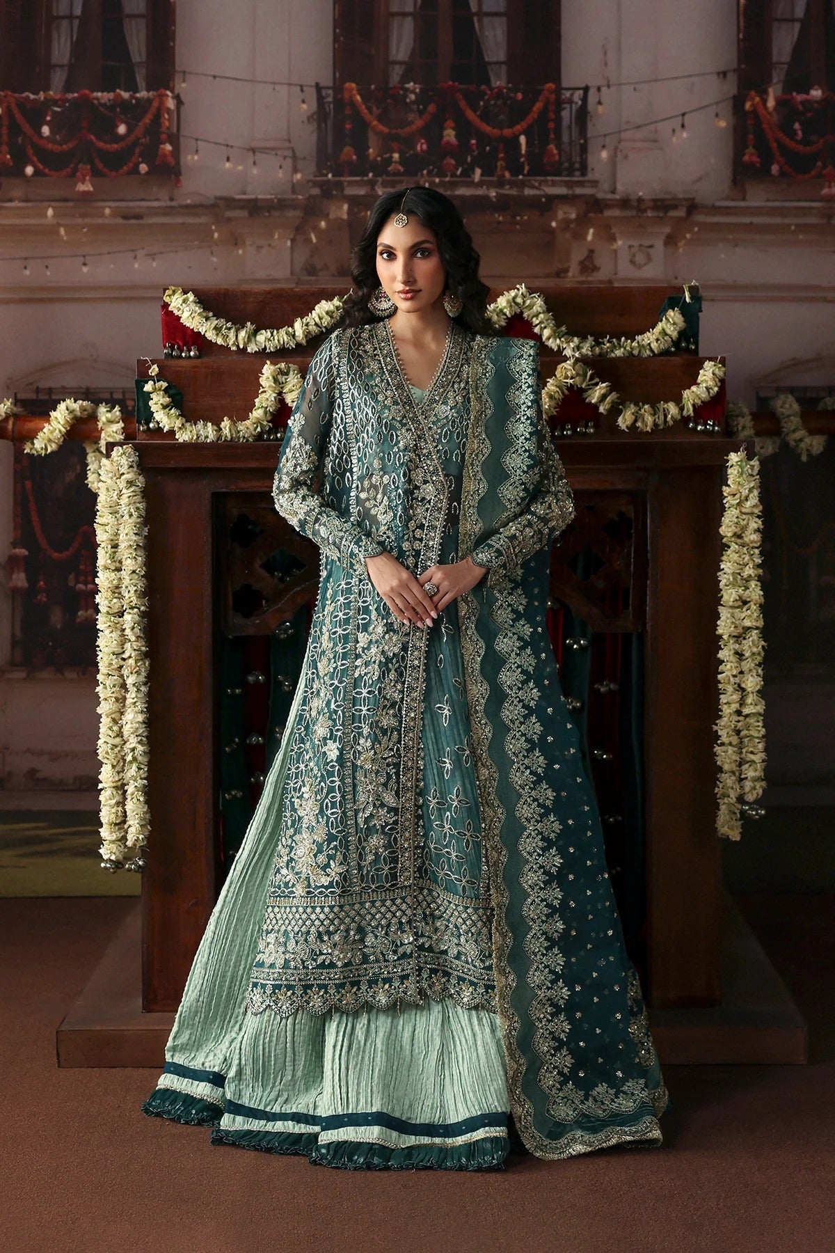A woman stands indoors wearing an ornate teal and mint green traditional outfit with heavy gold embroidery, a long sheer jacket, flared skirt, and matching dupatta. The setting features a wooden structure decorated with floral garlands and warm ambient lighting.