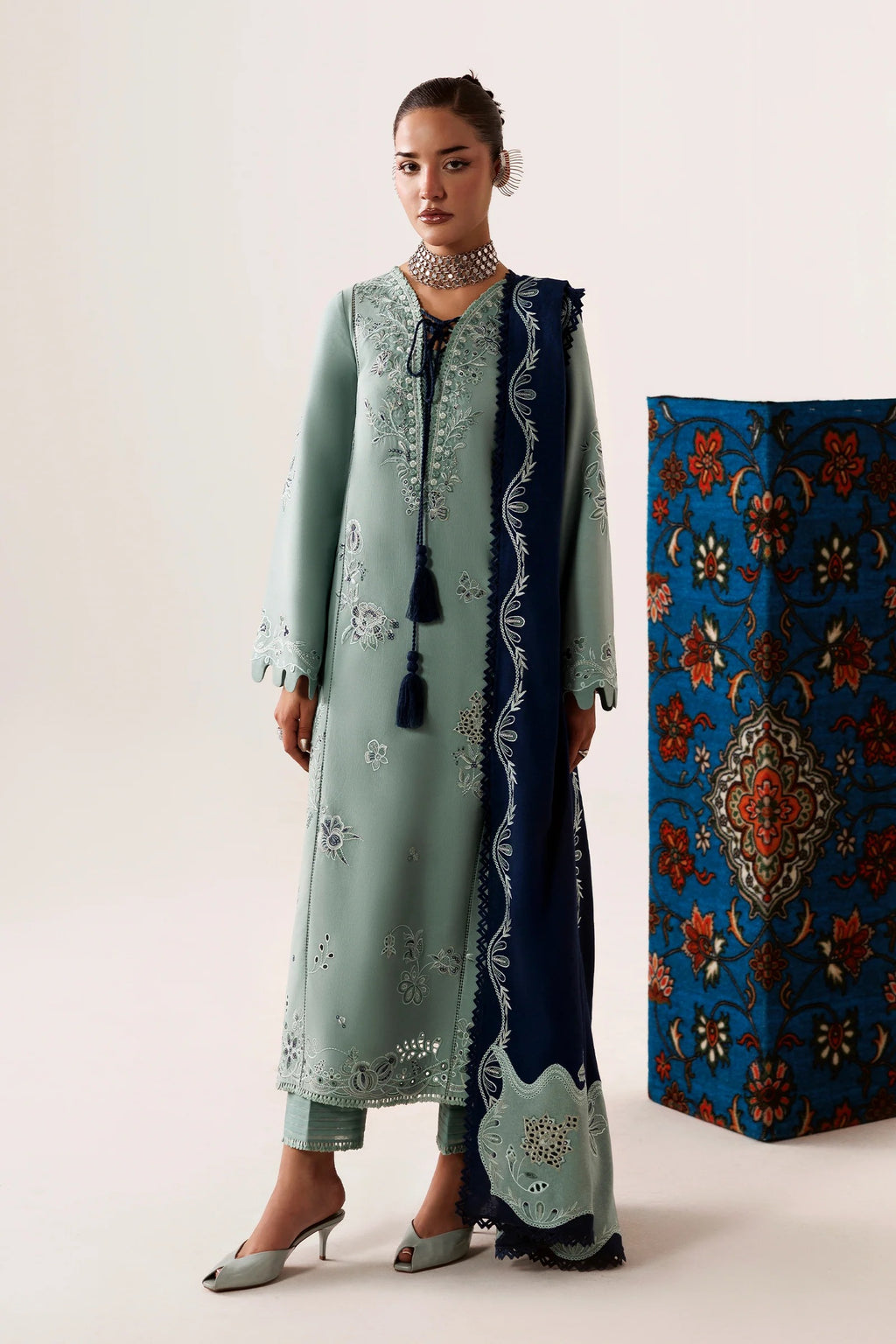A model wears a traditional three-piece salwar kameez suit in mint green and navy blue. The long kurta features silver mirror embroidery, paired with matching trousers and a navy blue dupatta with silver borders and tassels.