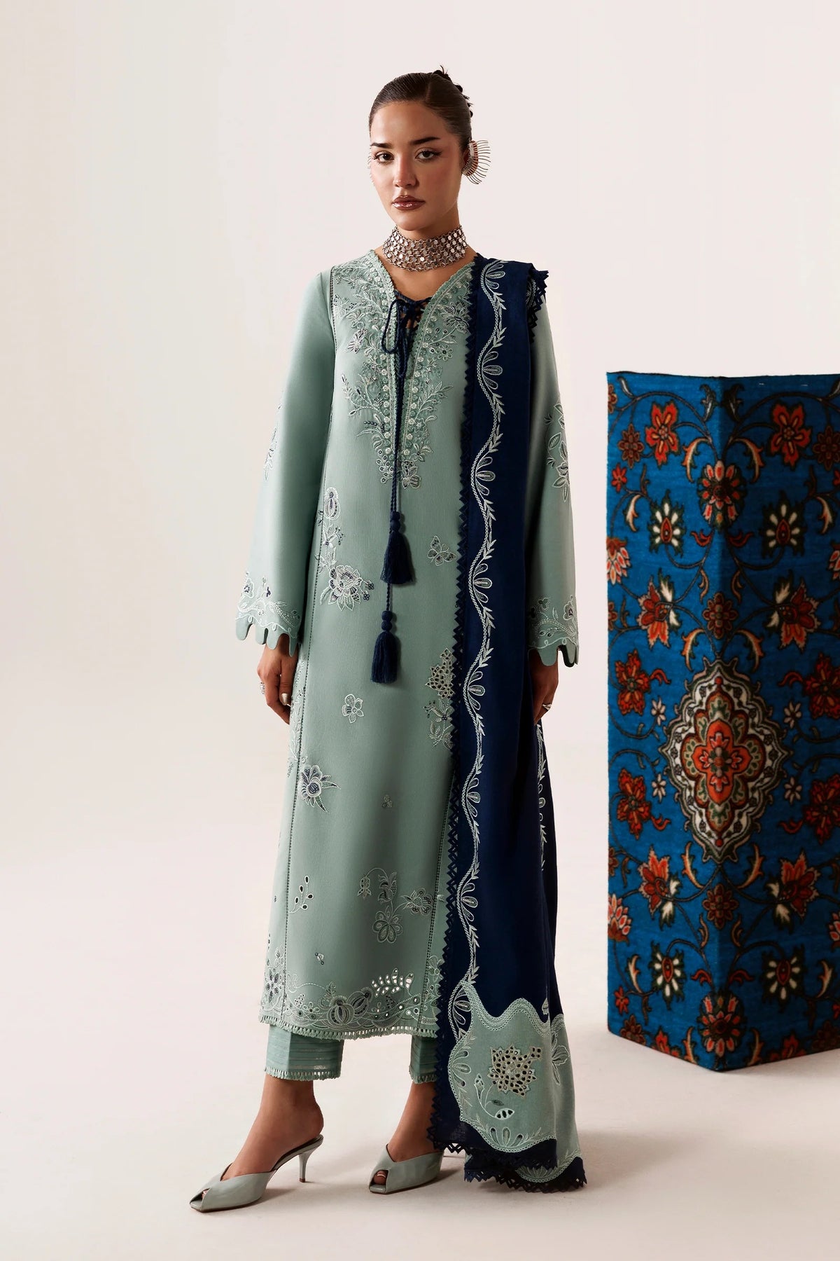 A model wears a traditional three-piece salwar kameez suit in mint green and navy blue. The long kurta features silver mirror embroidery, paired with matching trousers and a navy blue dupatta with silver borders and tassels.