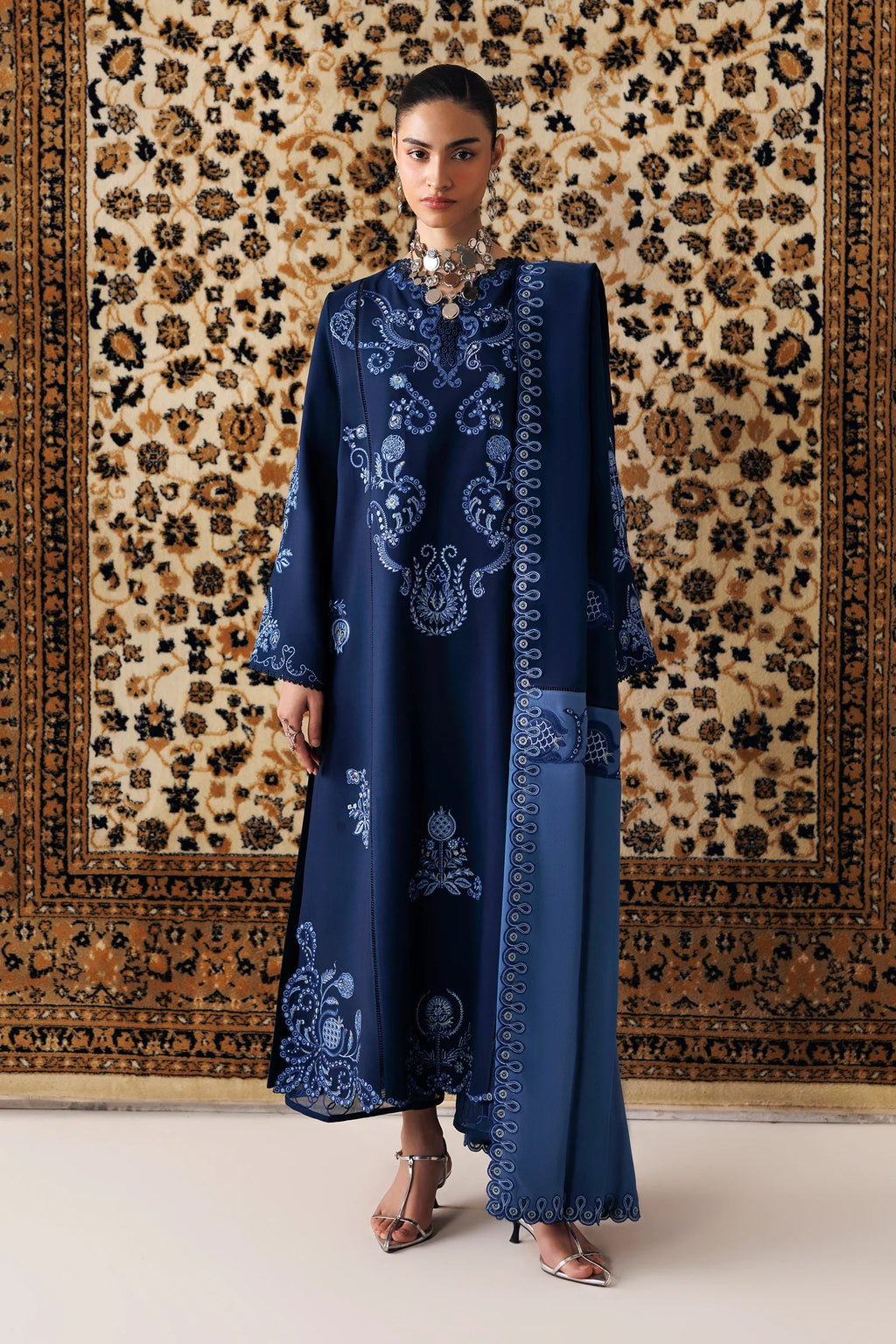 A woman models a long navy blue embroidered tunic with matching wide-leg trousers, standing in front of a large patterned tapestry.