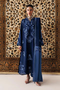 A woman models a long navy blue embroidered tunic with matching wide-leg trousers, standing in front of a large patterned tapestry.