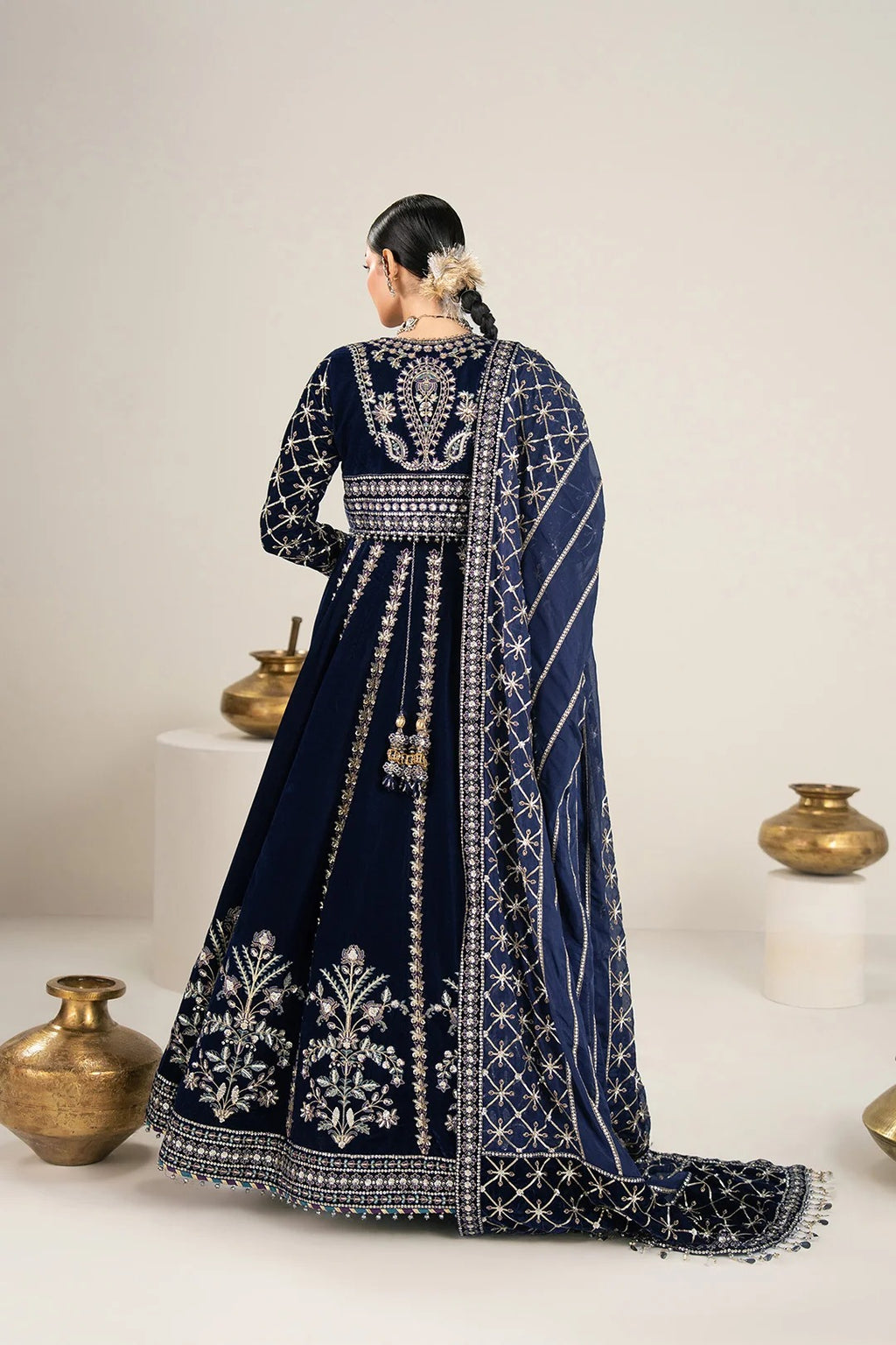 A woman wearing a navy blue traditional outfit with intricate silver embroidery, shown from the back in a studio setting with decorative gold vases on white pedestals.