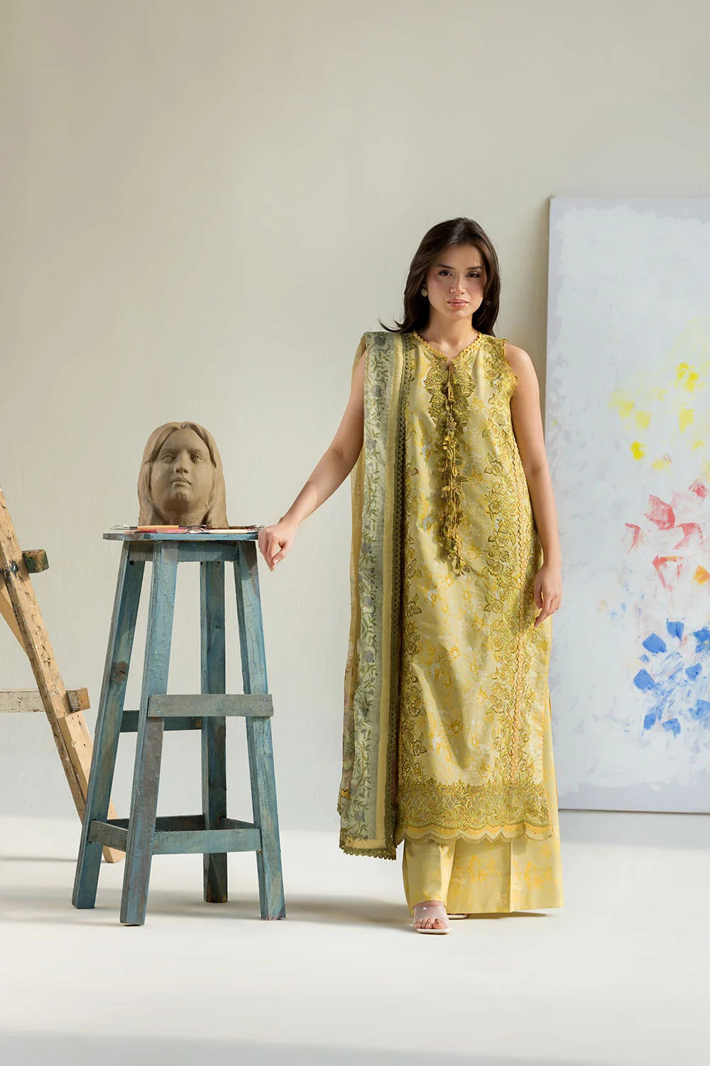 Woman in a yellow traditional outfit standing next to a wooden stool with a head on it, against a white wall with abstract art.