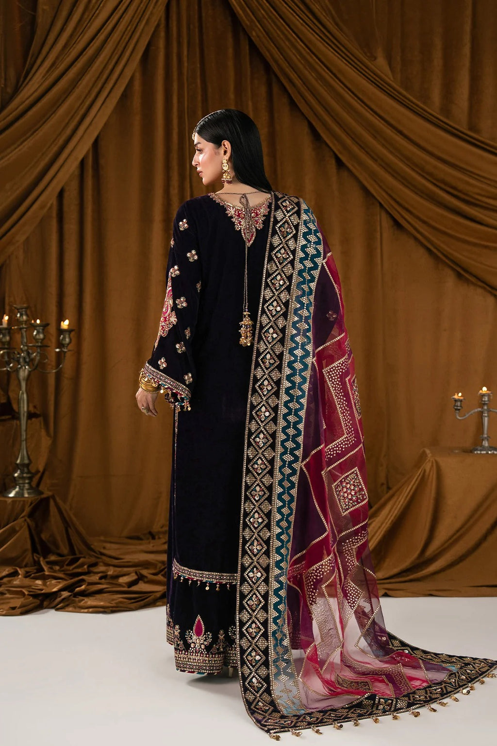A woman stands with her back to the camera wearing a traditional dark navy embroidered outfit with a pink, teal, and gold patterned dupatta, set against a draped brown fabric backdrop.