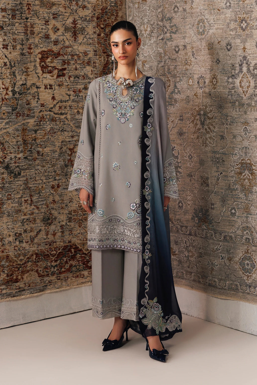 A woman models a traditional three-piece salwar kameez suit in a studio setting with a textured, vintage-style patterned rug. The set features a light gray kameez jacket with intricate silver and blue embroidery, matching gray trousers, and a dark navy blue dupatta with a gradient effect and detailed floral embroidery.