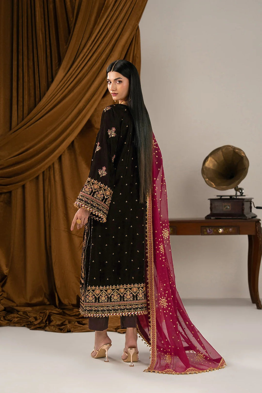 A woman modeling a black embroidered traditional outfit with gold detailing and a deep magenta dupatta with gold motifs, posed in front of a draped brown curtain and a vintage gramophone on a table.