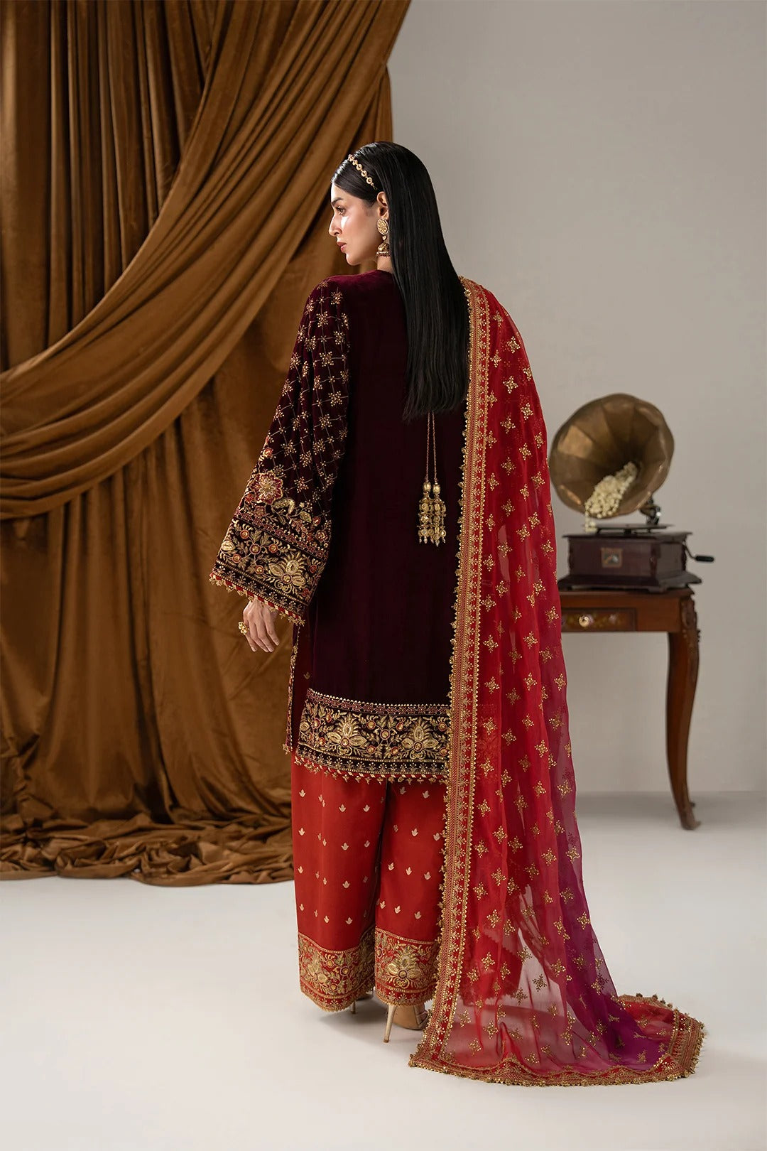 A woman stands in profile wearing a traditional South Asian outfit with a deep maroon velvet jacket, red patterned pants, and a red dupatta with gold embroidery, set against a studio backdrop with brown draped fabric and a vintage gramophone.