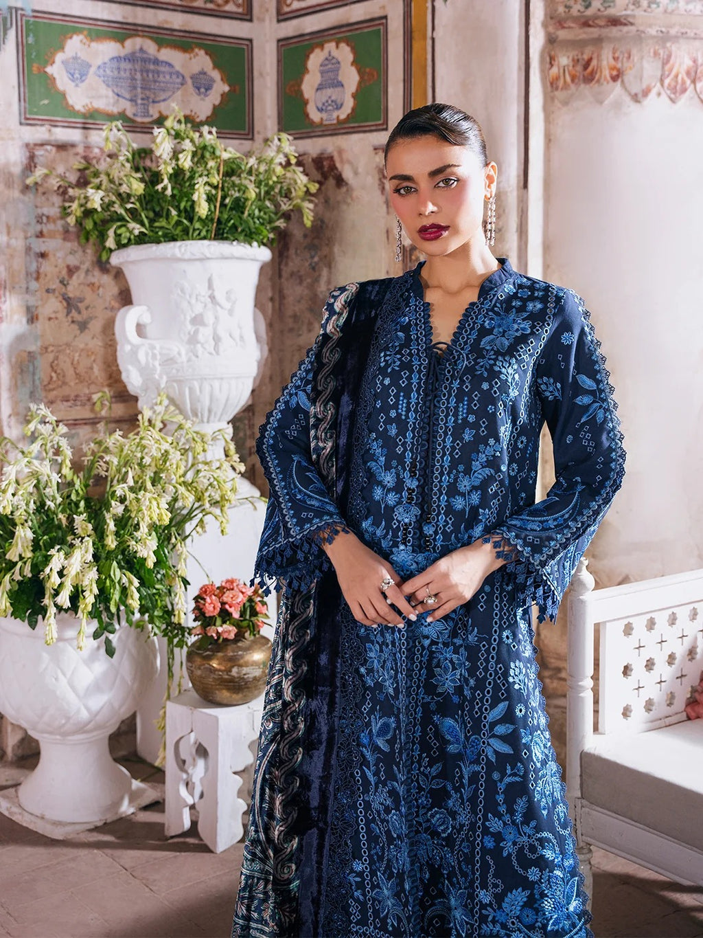 A woman models a dark blue embroidered traditional outfit with matching dupatta in an ornate indoor setting with classical wall panels, white vases, and potted plants.
