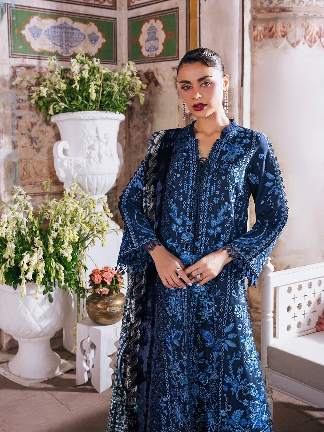 A woman models a dark blue embroidered traditional outfit with matching dupatta in an ornate indoor setting with classical wall panels, white vases, and potted plants.
