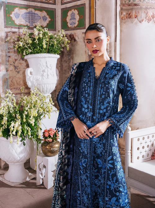 A woman models a dark blue embroidered traditional outfit with matching dupatta in an ornate indoor setting with classical wall panels, white vases, and potted plants.