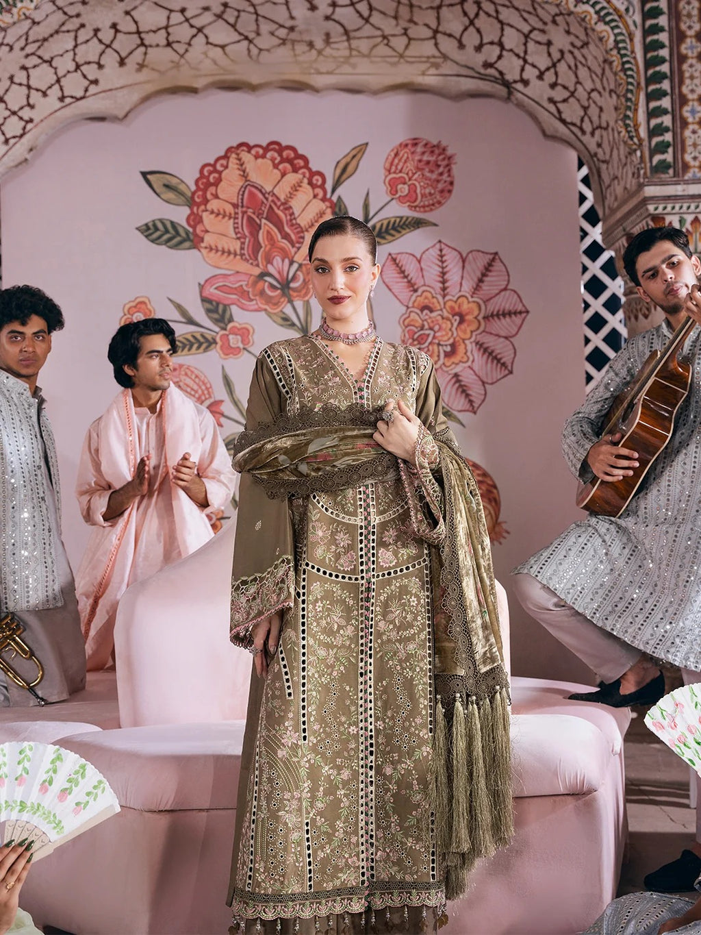 A woman stands in a pink decorated room wearing an ornate olive green embroidered traditional outfit with a matching dupatta. Two men sit on either side, one playing a guitar and the other clapping, with a large floral archway behind them.