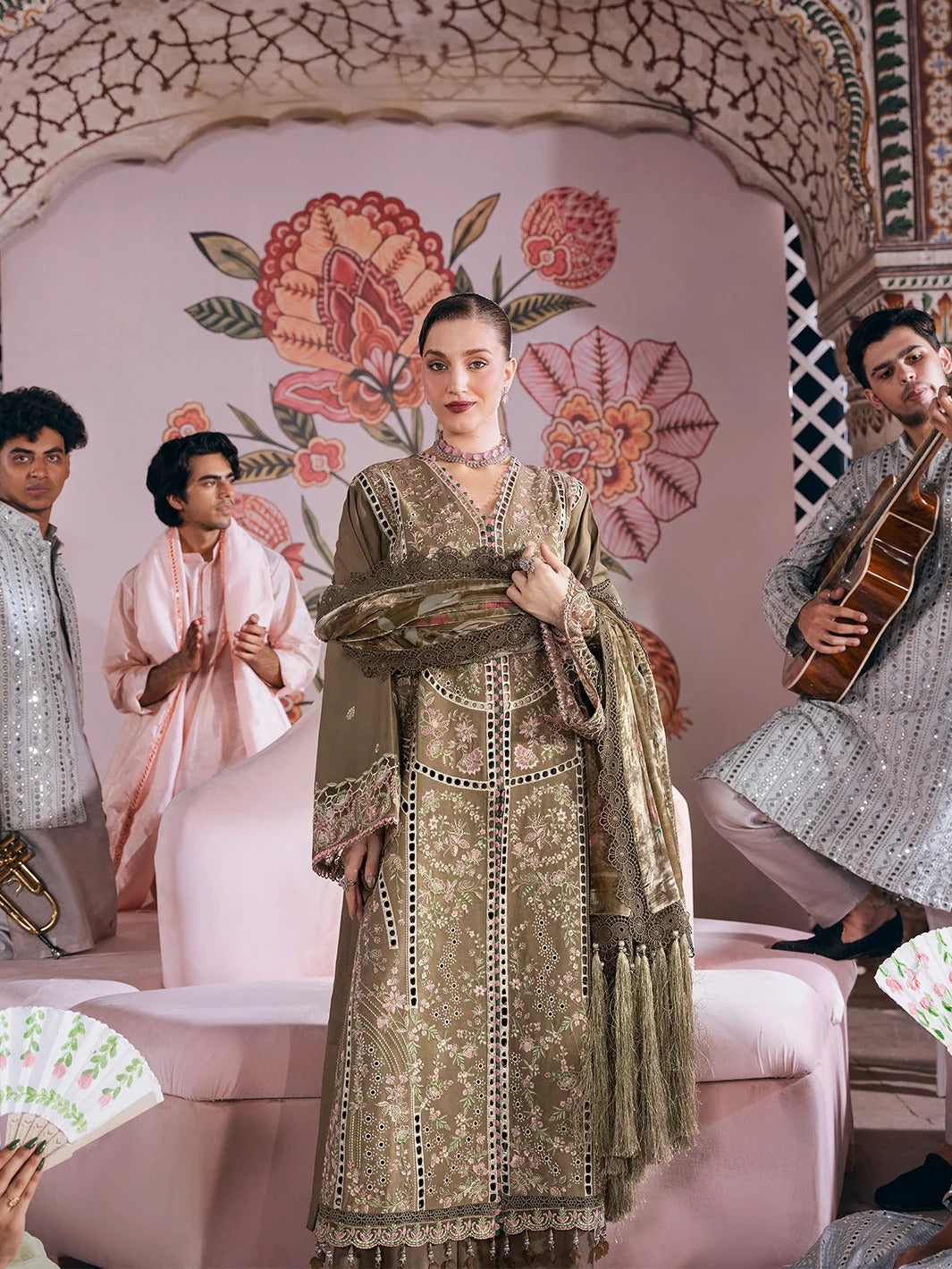 A woman stands in a pink decorated room wearing an ornate olive green embroidered traditional outfit with a matching dupatta. Two men sit on either side, one playing a guitar and the other clapping, with a large floral archway behind them.