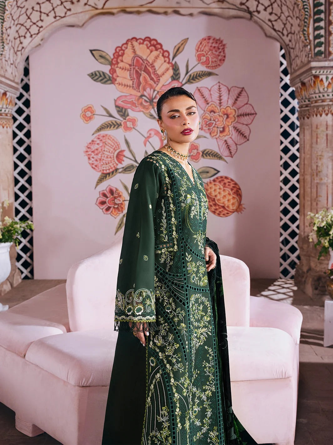 A woman models a dark green embroidered traditional dress with gold detailing, standing in front of a pink floral backdrop and a light pink sofa.