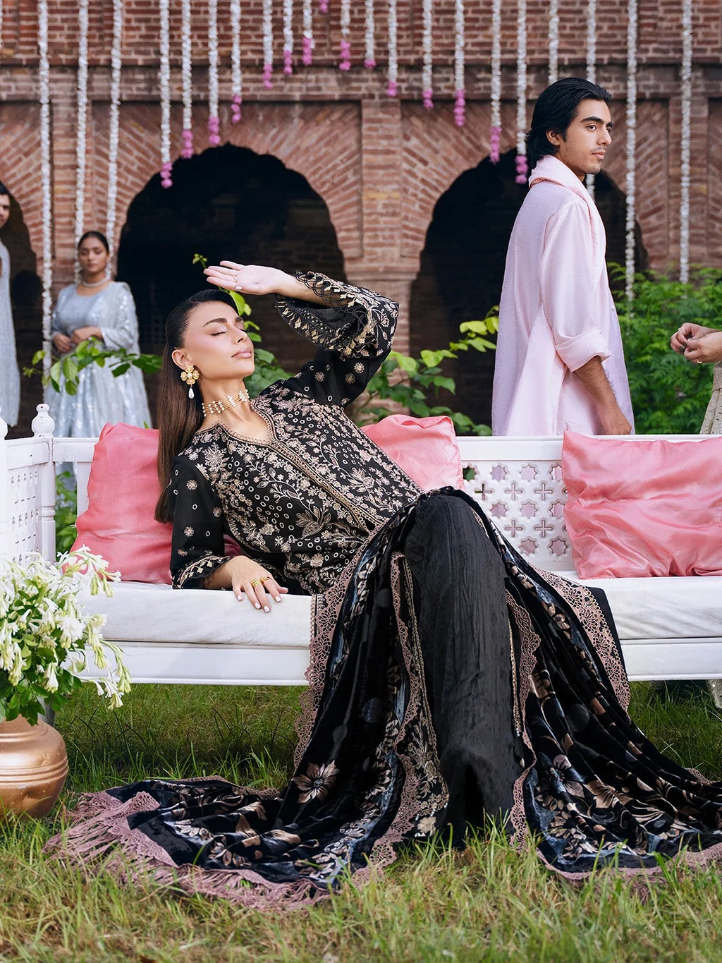A woman reclines on a white outdoor sofa wearing a black embroidered traditional outfit with metallic gold detailing and a matching draped scarf. A man in a light pink shirt stands nearby, with other people and a brick courtyard visible in the background.