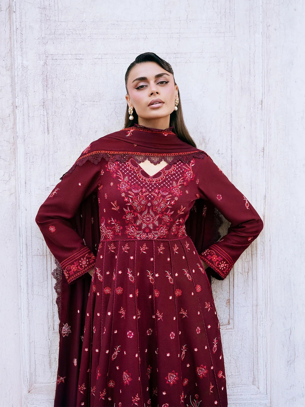 A woman wearing a deep maroon embroidered Anarkali gown with a matching dupatta, standing against a white textured wooden wall.