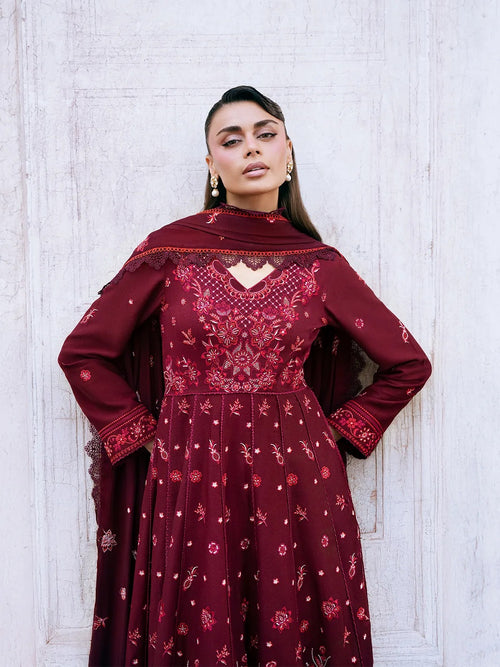 A woman wearing a deep maroon embroidered Anarkali gown with a matching dupatta, standing against a white textured wooden wall.