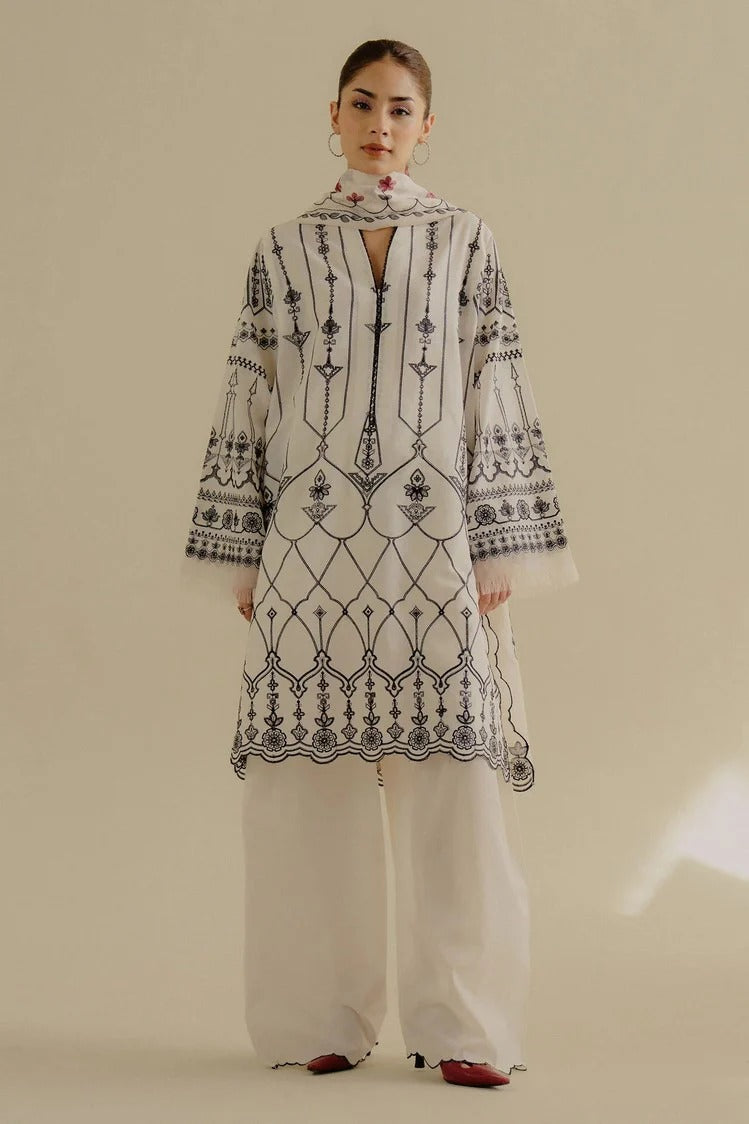 A model stands against a plain beige studio background wearing a white traditional outfit with extensive black geometric embroidery, wide-leg pants, and red pointed shoes.