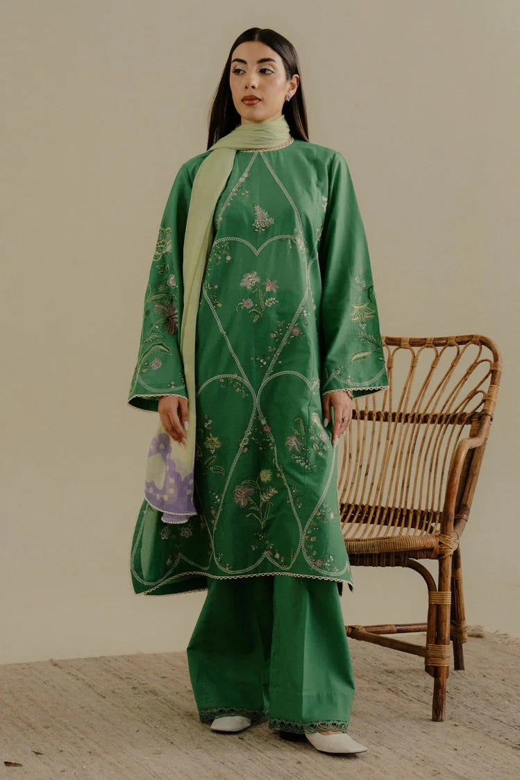 Woman wearing a green embroidered outfit with a beige wall and wicker chair in the background