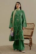 Woman wearing a green embroidered outfit with a beige wall and wicker chair in the background